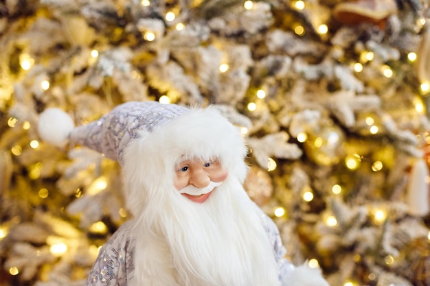 A toy of santa claus in a white suit stands on in living room near christmas tree Christmas tree with garlands of bokeh lights