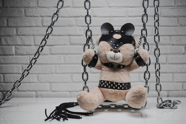 The toy Teddy bear dressed in leather belts and mask chained and handcuffed accessory for BDSM games on a light background texture of a brick wall