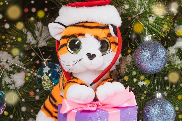 A toy tiger in a hat on the background of a Christmas tree with lights.