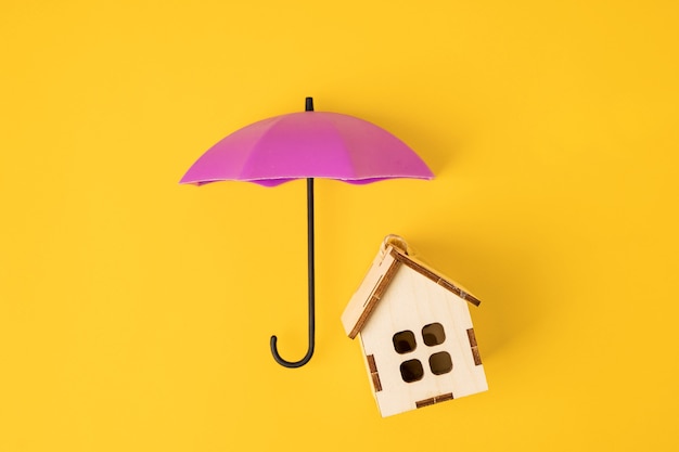 A toy umbrella over a wooden house on a yellow background. A symbol of care and reliability