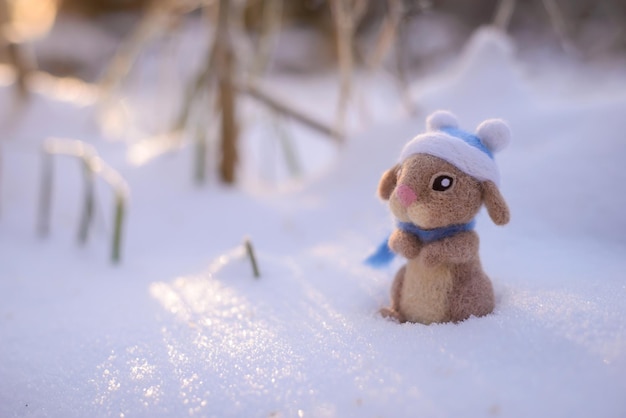 Toy wool felted cute rabbit, hare in a blue hat and scarf stands in the snow on a sunny winter day