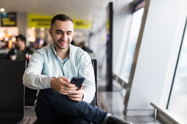TPortrait красавец в повседневной одежде, держа его багаж и сообщения через мобильный телефон, сидя в зале аэропорта