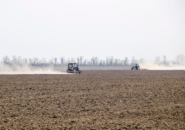 The tractor harrows the soil on the field and creates a cloud of dust behind it