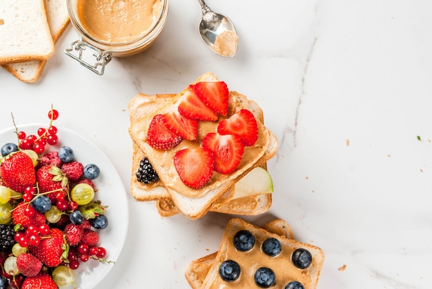 Traditional American and European summer breakfast: sandwiches of toast with peanut butter, berry, fruit apple, peach, blueberry, blueberry, strawberry, banana. White marble table. copyspace top view