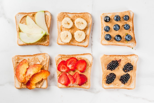 Traditional American and European summer breakfast: sandwiches of toast with peanut butter, berry, fruit apple, peach, blueberry, blueberry, strawberry, banana. White marble table. copyspace top view