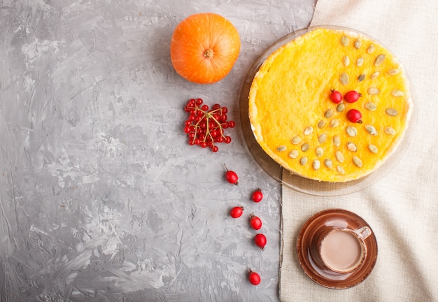 Traditional american sweet pumpkin pie decorated with hawthorn red berries and pumpkin seeds with cup of coffee on a gray concrete background 