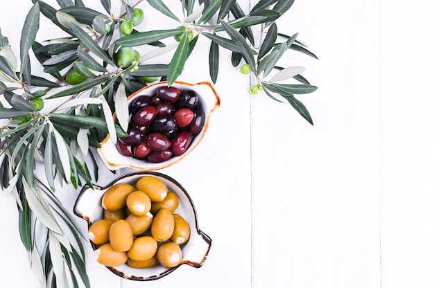 Traditional appetizers, green and red olives from Greek cuisine. White wood background. Fresh branches of olives. Copy space