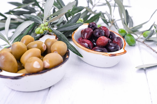 Traditional appetizers, green and red olives from Greek cuisine. White wood background. Fresh branches of olives. Copy space