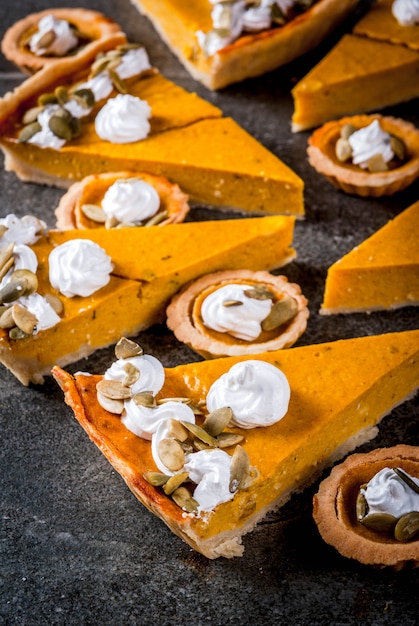 Traditional autumn dishes. Halloween, Thanksgiving. Set of spicy pumpkin pie and pumpkin tartlets with whipped cream & pumpkin seeds on black stone table. Copy space