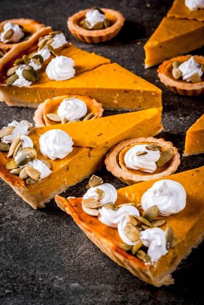 伝統的な秋の料理。ハロウィーン、感謝祭。スパイシーなカボチャのパイと黒い石のテーブルにホイップクリームとカボチャの種とカボチャのタルトのセット。コピースペース