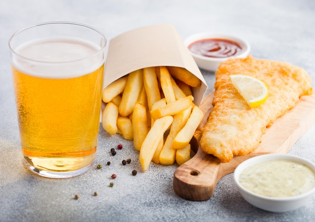 Traditional British Fish and Chips with tartar sauce abd glass of craft lager beer and tomato ketchup on chopping board