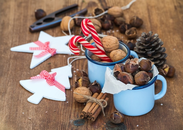 Traditional Christmas foods and decoration. Roasted chestnuts in blue  enamel mug, walnuts, cinnamon sticks, candy canes, pine cone on rustic wooden 