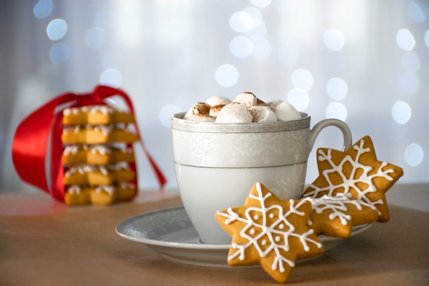 Традиционные рождественские пряники ручной работы и чашка горячего зимнего напитка с зефиром, стогом печенья и боке на фоне.