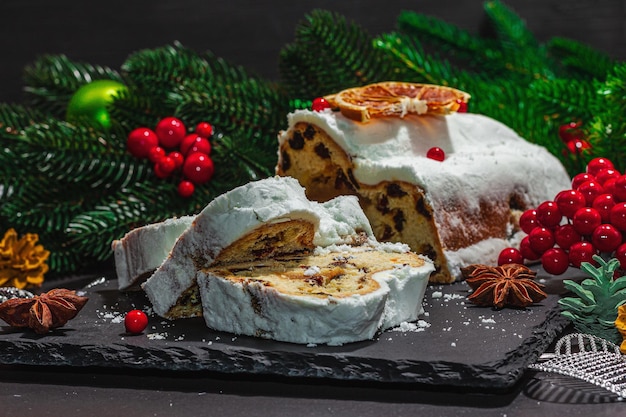 Traditional Christmas stollen German cake European pastry fragrant home baked bread with spices and dried fruit Xmas tree branches and decorations black background close up