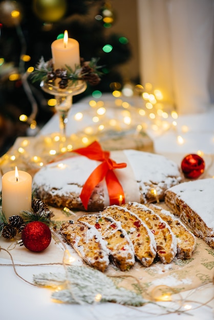 Traditional Christmas stollen made of dried fruits and nuts sprinkled with powdered sugar on the background of a Christmas decor with candles. Traditional Christmas cupcake.