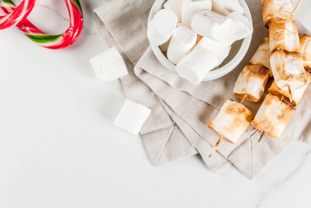 Traditional christmas sweets  candy cane, marshmallow and baked on fire marshmallow skewers on white , copyspace top view