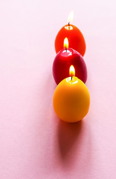 Traditional Easter decor. Group of bright burning paraffin candles in the shape of colorful eggs on pink background.