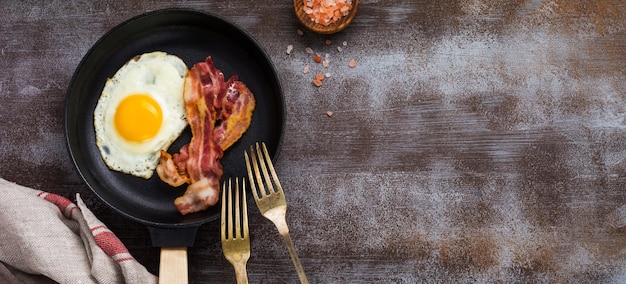 Traditional English breakfast with fried eggs and bacon in cast iron pan on dark concrete background. Top view.