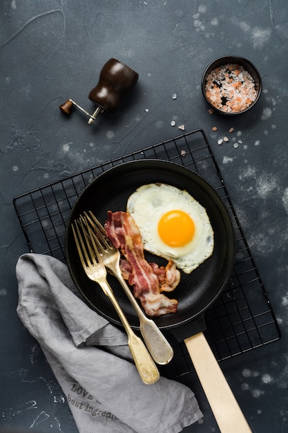 Traditional English breakfast with fried eggs and bacon in cast iron pan on dark concrete background. Top view.