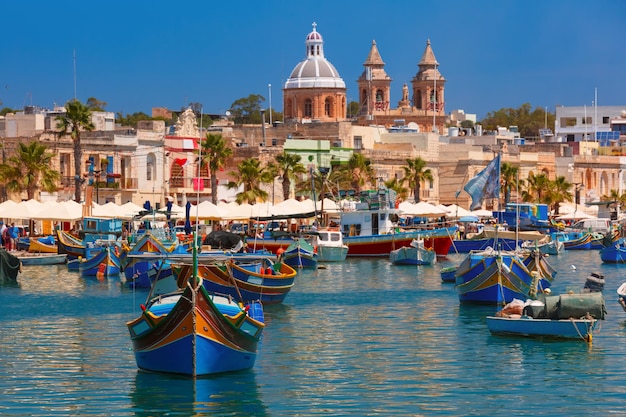 Traditional eyed colorful boats Luzzu and Parish Church of Our Lady of Pompei in the Harbor of Mediterranean fishing village Marsaxlokk, Malta