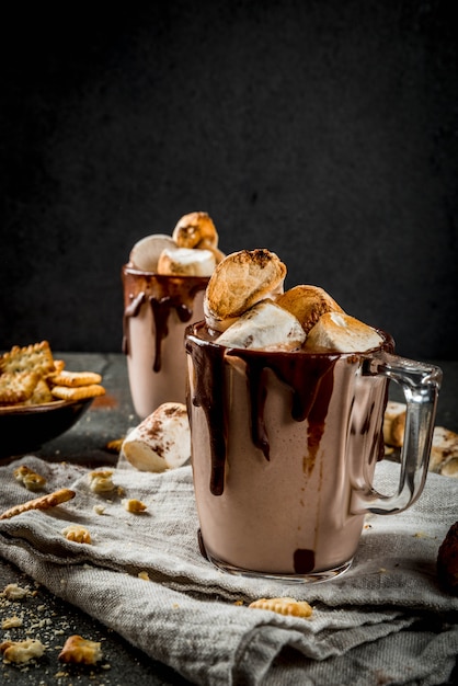 Traditional fall and winter cocktails, alcohol. Campfire hot chocolate eggnog cocktail with salted crackers and roasted marshmallow, in two mugs, on black stone table, 