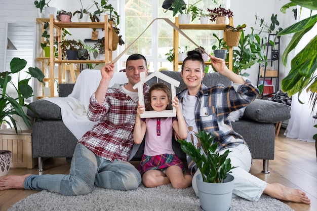 子供を持つ伝統的な家族は、ソファに座って屋根を持って新しい家を楽しんでいます 住宅ローン保険と保護を購入し、鉢植えの植物のある自分の家の温室に引っ越します