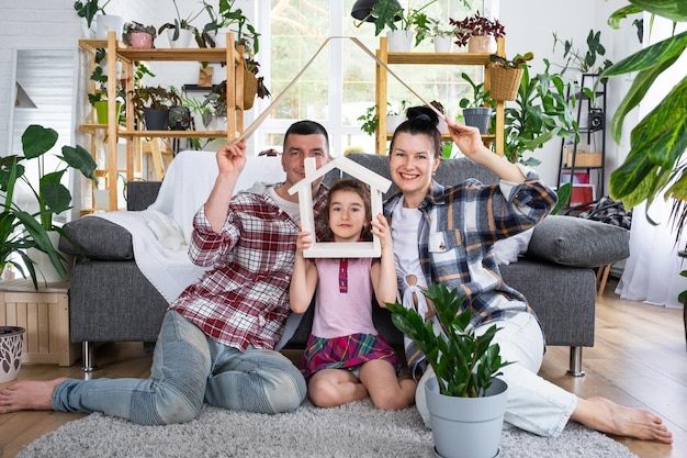 Традиционная семья с ребенком наслаждается новым домом, сидя на диване и держась за крышу. Страхование ипотеки и защита покупают и переезжают в собственный домашний зеленый домик с горшечными растениями.