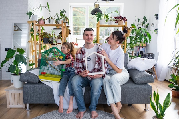 Foto una famiglia tradizionale con un bambino e un pappagallo in gabbia si gode una nuova casa seduta sul divano e tiene il tetto acquistando un'assicurazione ipotecaria e traslocando nella propria casa