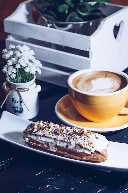 Traditional french dessert eclair with white chocolate coffee\
cup and white box with plants in a cafe or restaurant