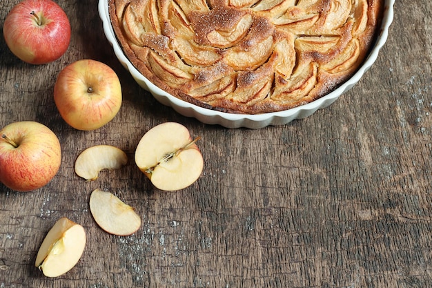 Traditional french homemade apple pie in a white ceramic baking form and apples on wooden table