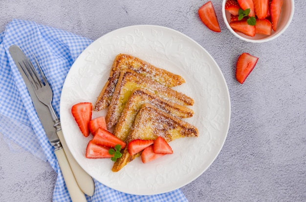 Traditional French toast sprinkled with powdered sugar with fresh strawberries on a white plate with a cup of tea