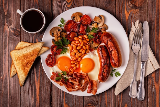 Traditional full english breakfast with fried eggs, sausages, beans, mushrooms, grilled tomatoes and bacon on wooden background