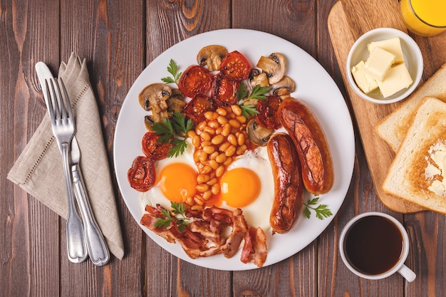 Традиционный полный английский завтрак с яичницей, сосисками, фасолью, грибами, жареными помидорами и беконом на деревянном фоне