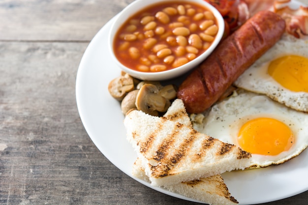 Traditional full English breakfast with fried eggs, sausages, beans, mushrooms, grilled tomatoes and bacon on wooden surface