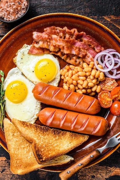 Традиционный полный английский завтрак с яичницей, сосисками, беконом, фасолью и тостами. Темный деревянный фон. вид сверху.