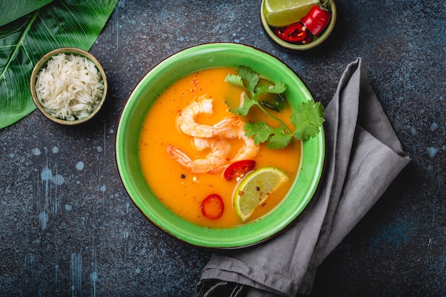 Traditional healthy Thai soup tom yum with shrimps, lime, coriander in bowl on rustic background with white rice, overhead shot. Authentic Thai food concept