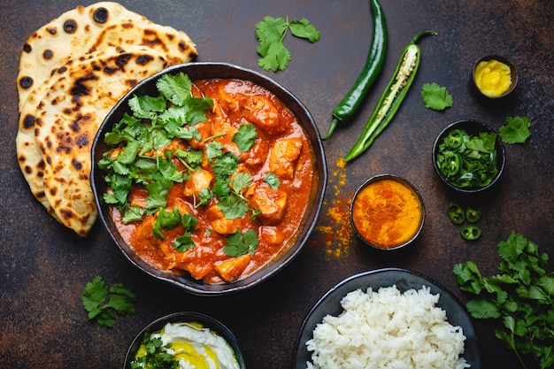 Traditional Indian dish Chicken tikka masala with spicy curry meat in bowl, basmati rice, bread naan, yoghurt raita sauce on rustic dark background, top view, close up. Indian style dinner from above