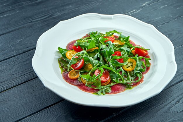 The traditional Italian appetizer salad is vitello tonnato. Thinly sliced beef with rucola, tomatoes and pesto, served in a white plate on a dark surface