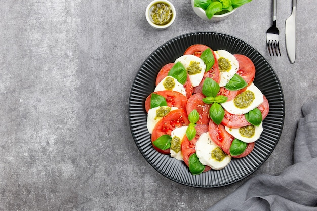 Traditional italian caprese salad with sliced tomatoes, mozzarella, basil, pesto sauce and spice on a dark gray concrete background. Top view. Copy space.
