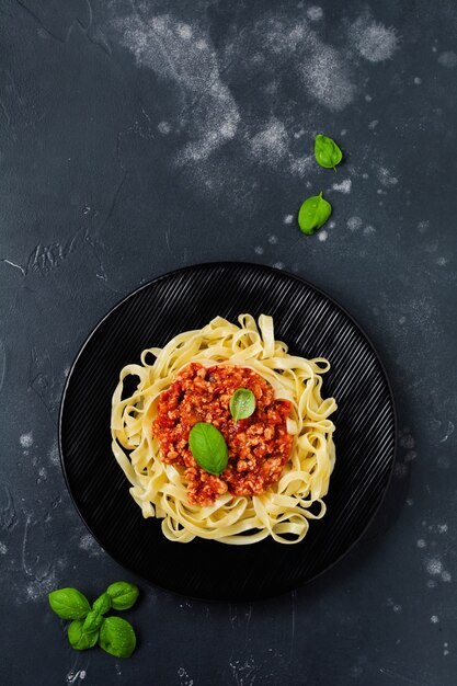 Traditional italian dish fettuccine pasta with bolognese sauce, basil and parmesan cheese in black plate on dark wooden surface. top view