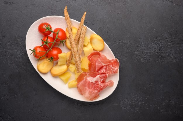 Traditional Italian food is grissini bread with prosciutto, cheese and tomatoes with herbs on a plate on a dark background.