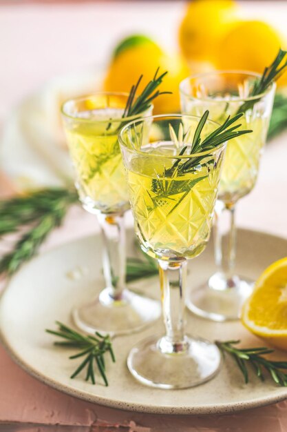 Traditional italian homemade lemon alcohol drink liqueur limoncello with pieces of lemon and rosemary herb on light pink peach or coral color stone concrete surface