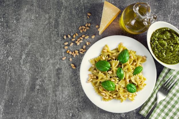 Traditional italian pasta with fresh vegetables, parmesan cheese, basil leaves, pine nuts and pesto sauce in white plate on grey stone background. Top view, flat lay, copy space.