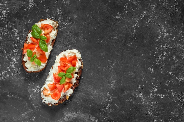 Traditional Italian snack. Bruschetta caprese toast with tomatoes, soft cottage cheese and basil on a dark black background.