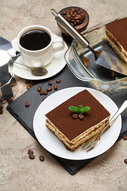 Traditional italian tiramisu dessert in glass baking dish and portion on grey concrete table