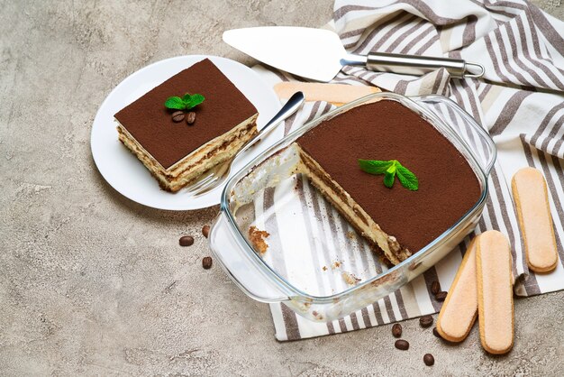 Traditional italian tiramisu dessert in glass baking dish savoiardi cookies and portion on grey concrete table