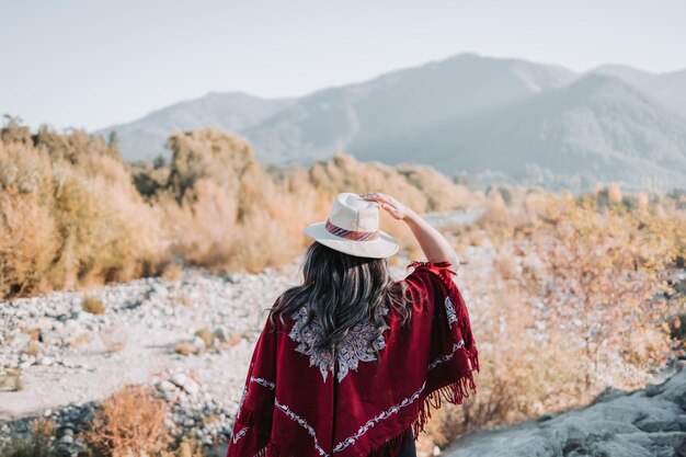 Traditional latin american woman wearing a red poncho and a hat seen from behind in a natural space