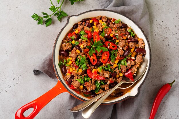 Traditional Mexican food - Chili con carne with minced meat and vegetables stew in tomato sauce in a cast iron pan on light gray slate or concrete background. Top view with copy space