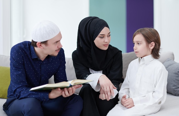 Родители традиционной мусульманской семьи с детьми читают Коран и вместе молятся на диване перед ужином ифтар во время праздника рамадан дома