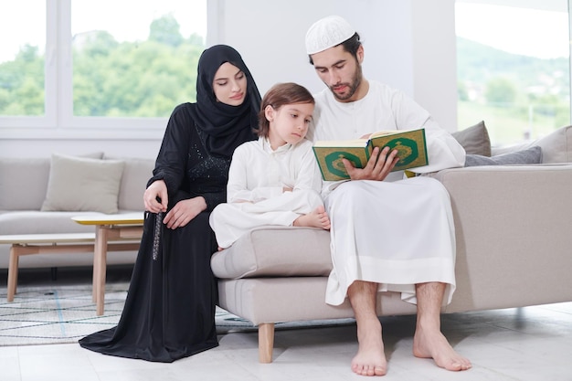 Traditional muslim family parents with children reading Quran and praying together on the sofa before iftar dinner during a ramadan feast at home